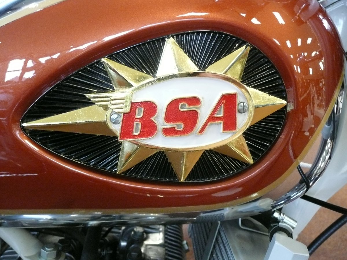 1960s BSA star tank badge: a gold star behind a cream oval with a red winged BSA, over a black sunburst, on a copper tank