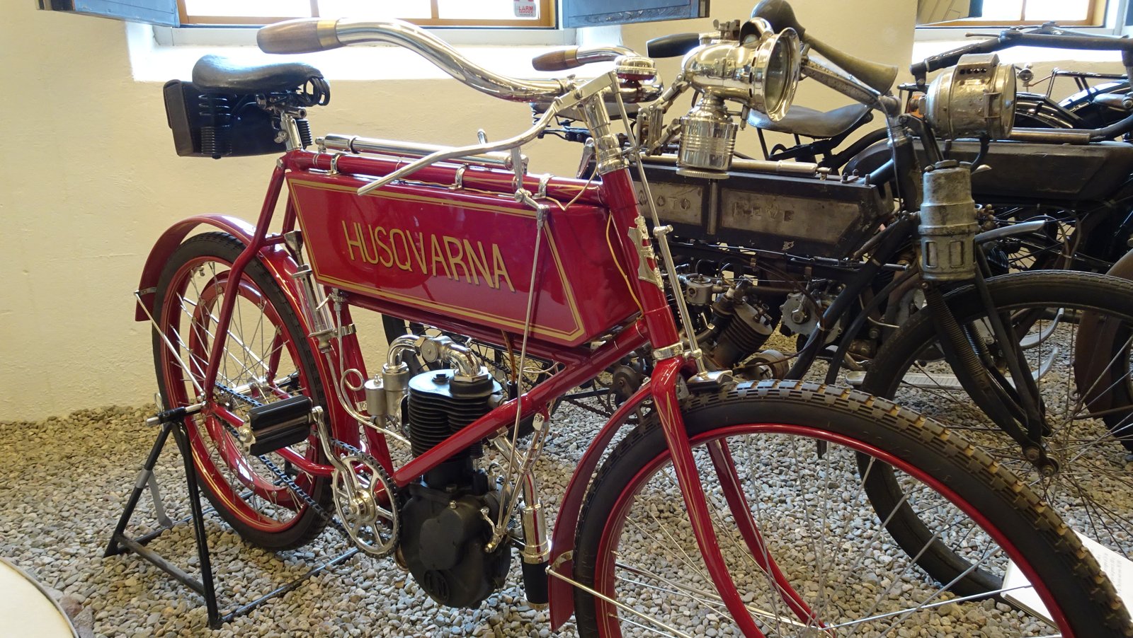 Red 1905 Husqvarna 300 cc motorcycle with HUSQVARNA in gold serif capitals on a lined red tank panel