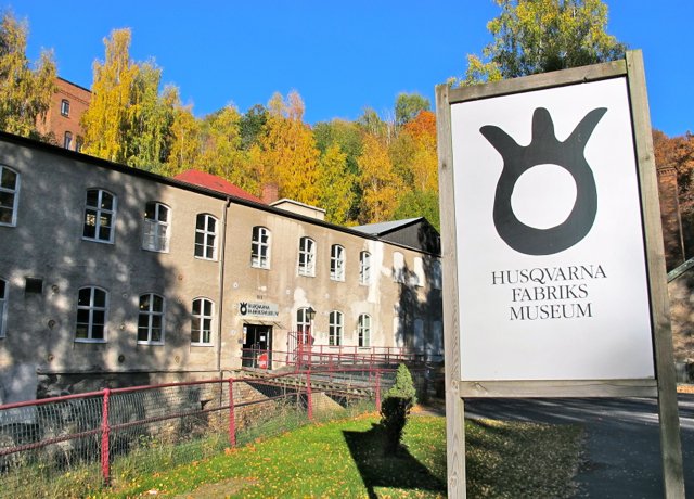 Sign for the Husqvarna Fabriksmuseum showing a black ring with three prongs above the museum's name, in front of the old factory building