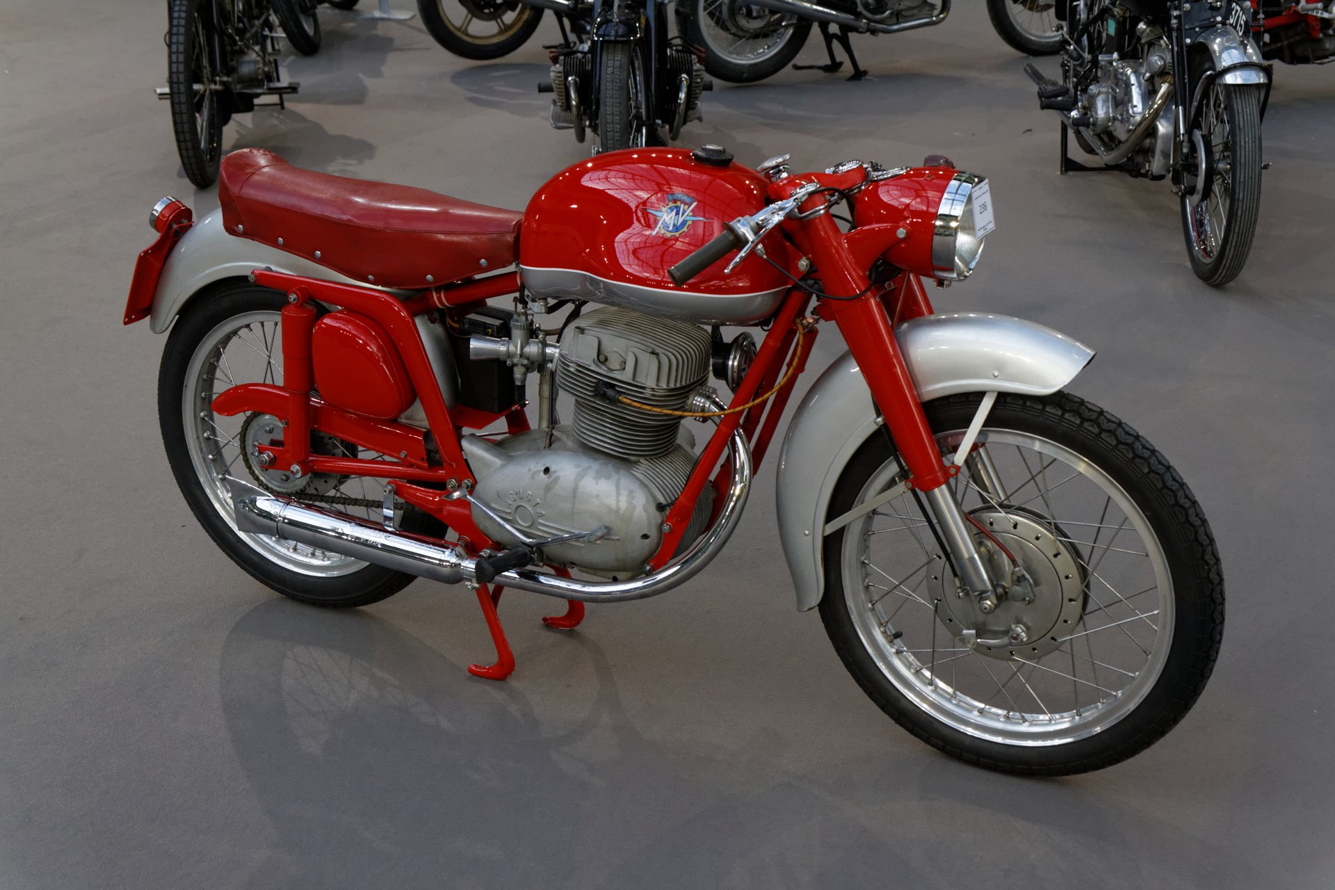 A red 1955 MV Agusta 175 CS Disco Volante with a bulbous red tank carrying the AGUSTA crest, in an auction hall
