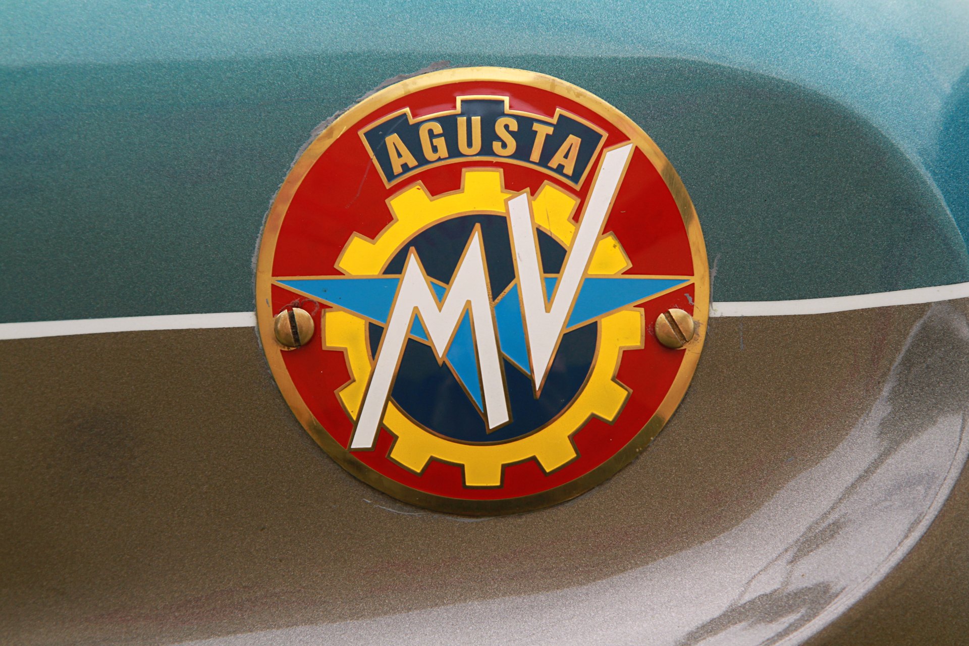 Close-up of a round enamel MV Agusta tank badge fixed with two brass screws: the crest on a red field inside a brass rim, with a yellow cog, navy centre, pale blue wings, white MV and a navy AGUSTA banner