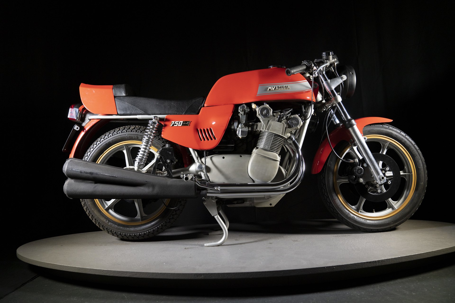 A red 1975 MV Agusta 750 S America on a turntable in a dark studio, with a chrome MVAGUSTA badge on its angular tank