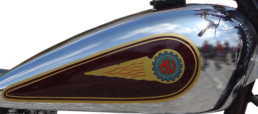 Chrome fuel tank of an MV Agusta 98 with a maroon panel edged in gold, containing a gold feathered wing that ends in a blue cog enclosing a red MV monogram