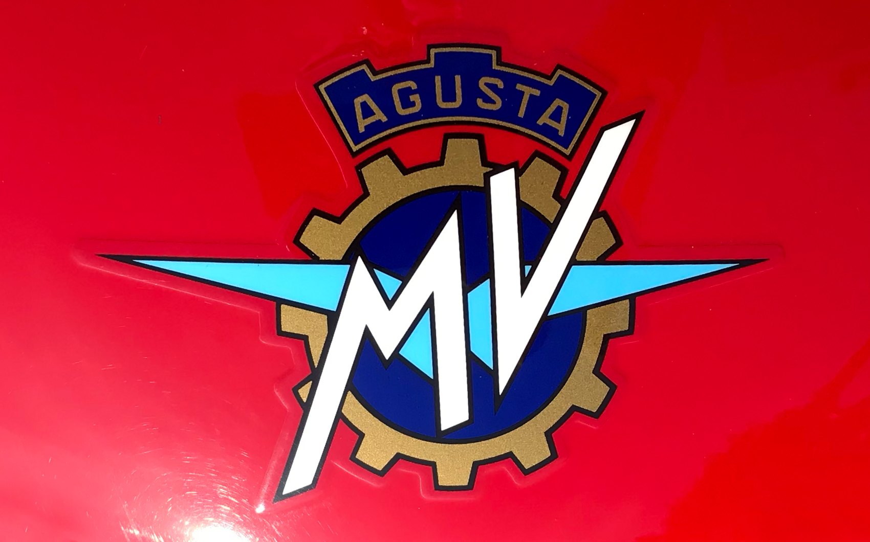 Close-up of the MV Agusta crest decal on the red tank of an F3: blue AGUSTA banner, bronze-gold cog, navy centre, pale blue wings and a white MV