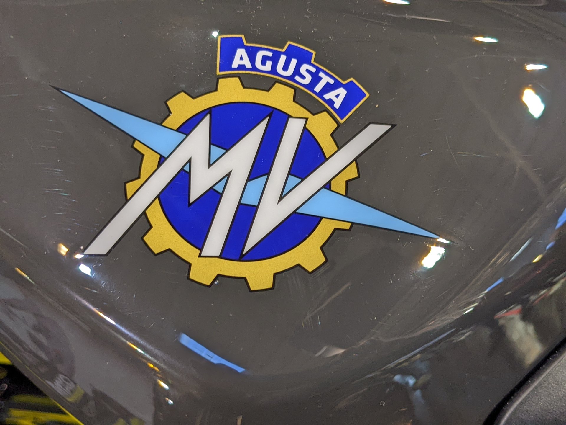 Close-up of the MV Agusta crest on the dark grey tank of a Rush: bright blue AGUSTA banner, yellow-gold cog, bright blue centre, pale blue wings and a white MV