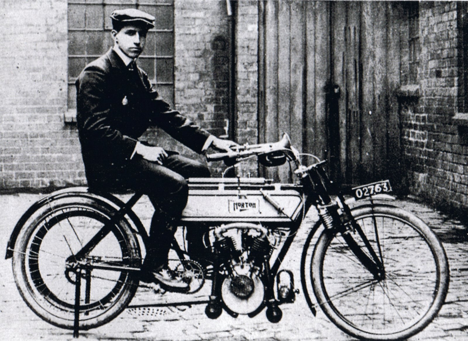 Black and white photograph of Rem Fowler in a flat cap sitting on the 1907 Norton twin, with NORTON lettered in a small panel on the side of the tank