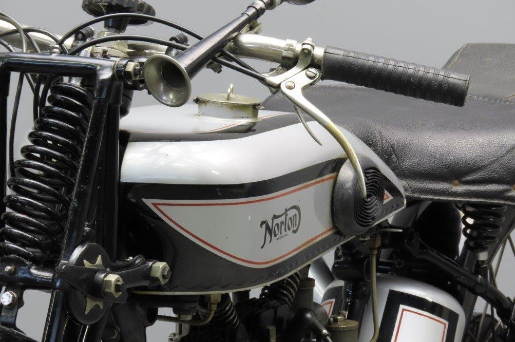 Silver saddle tank of a 1929 Norton Model 19 with a black curly N Norton transfer inside a panel lined in black and red