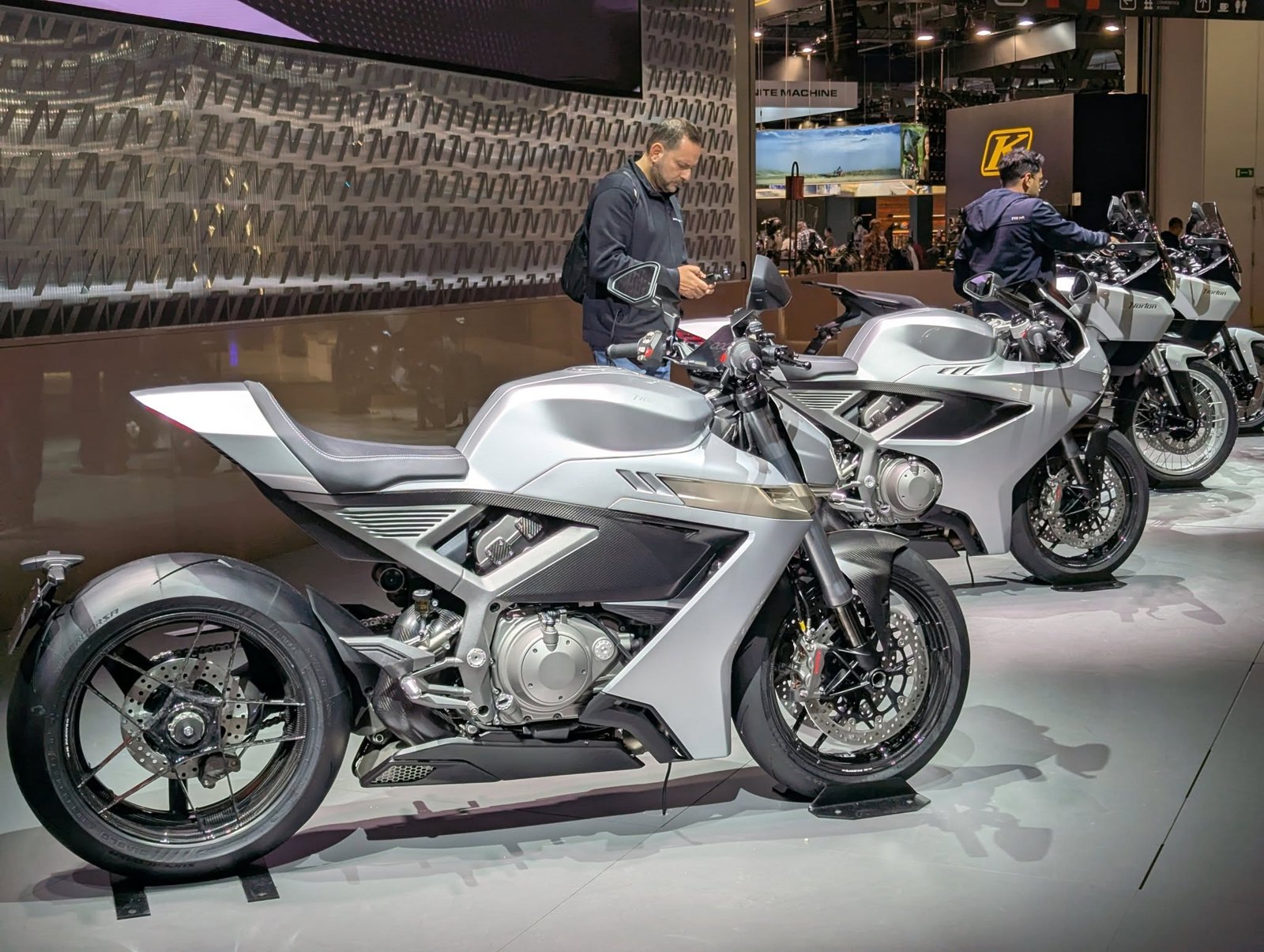 A silver 2026 Norton Manx on the EICMA 2025 stand, in front of a wall patterned with a repeated angular N