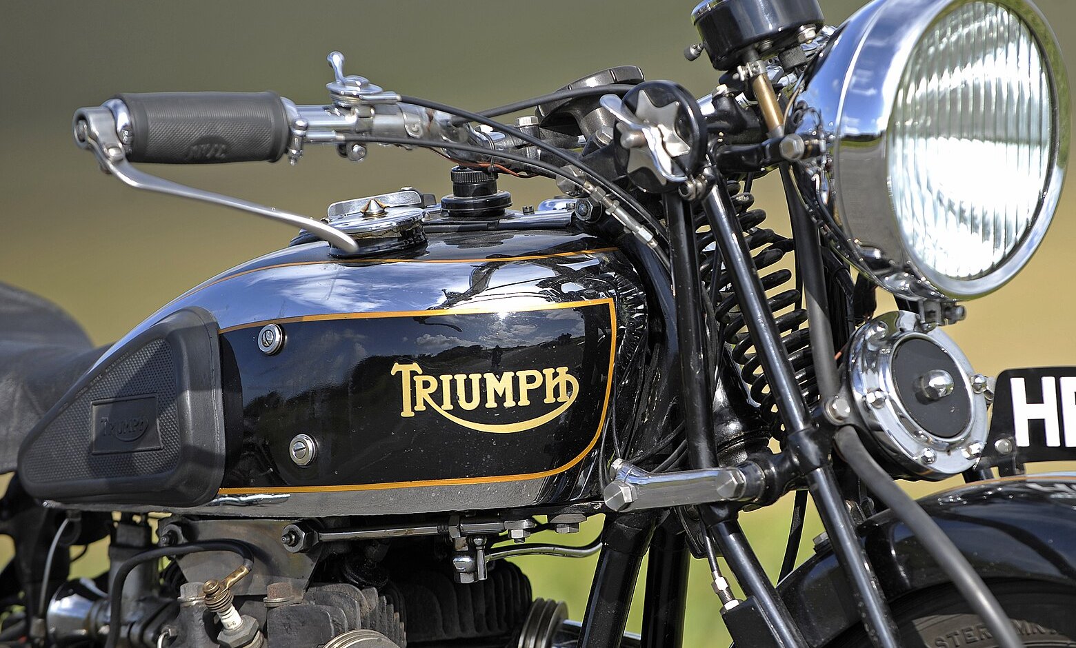 Black tank of a Triumph 6/1 with a gold swash TRIUMPH wordmark inside a gold-lined panel, with the headlamp and handlebars