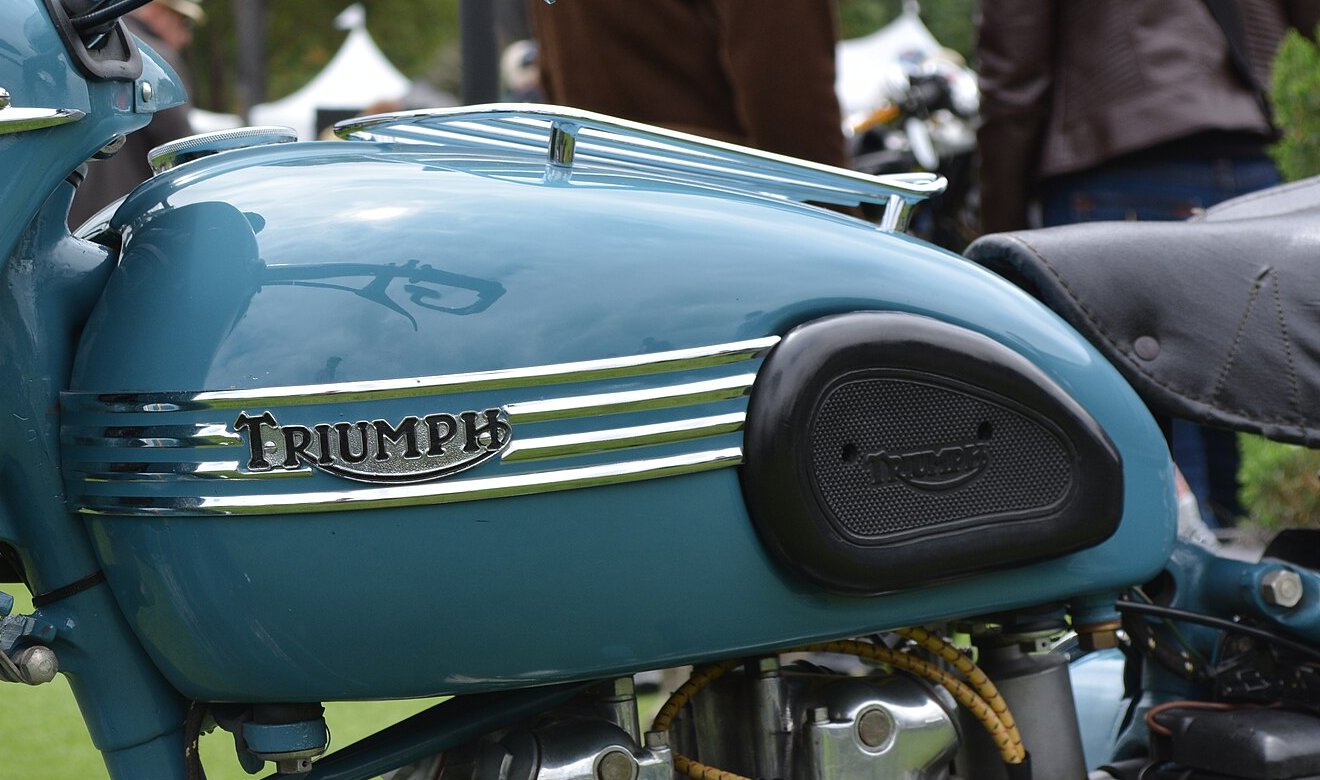 Pale blue 1950s Triumph tank with a chrome TRIUMPH script plate flanked by horizontal chrome bands, a rubber knee pad and a tank-top parcel grid