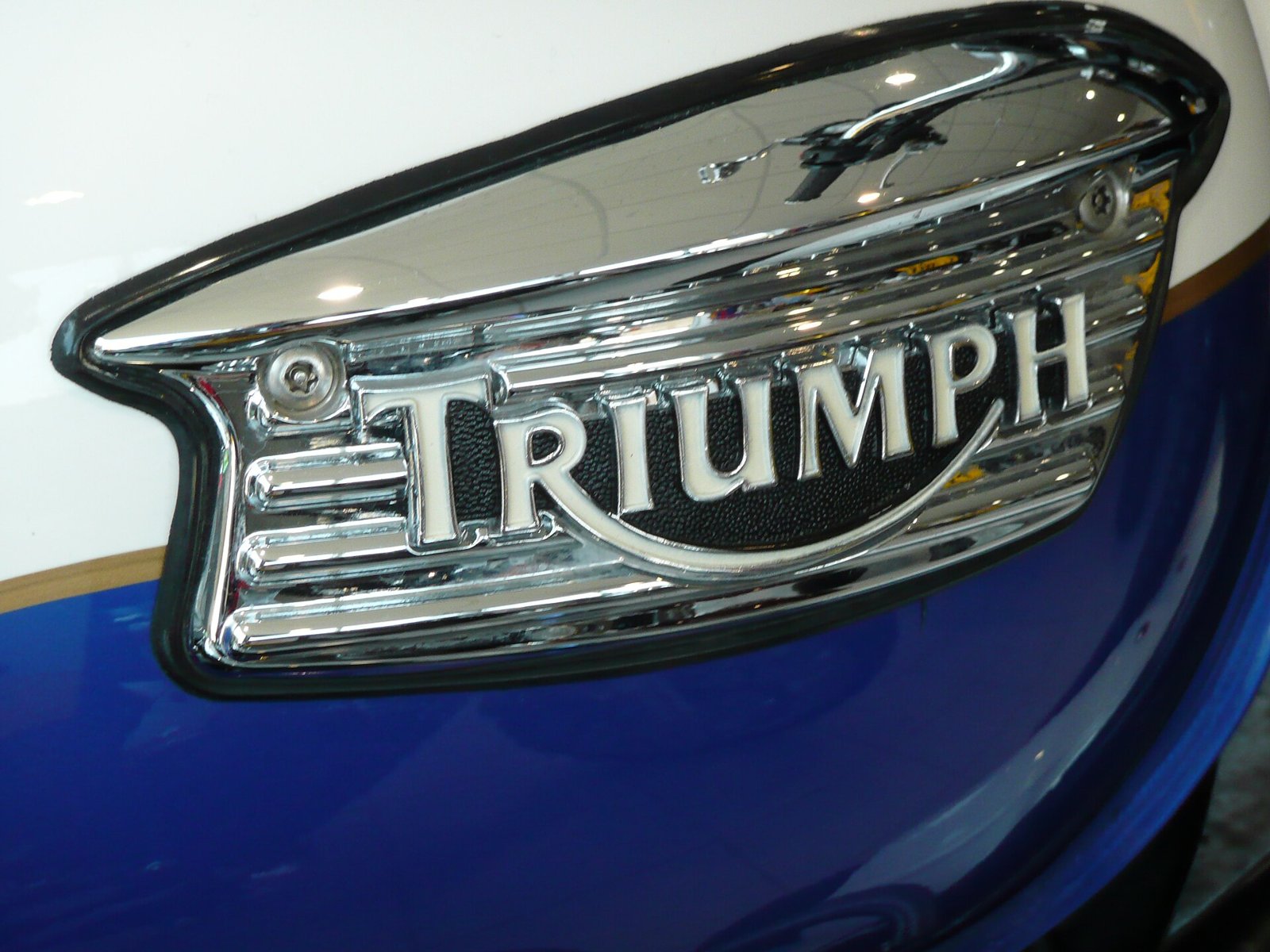 Close-up of a chrome Triumph tank badge of 2008: the wordmark with smile line on a black field, set in a chrome surround with horizontal ribs