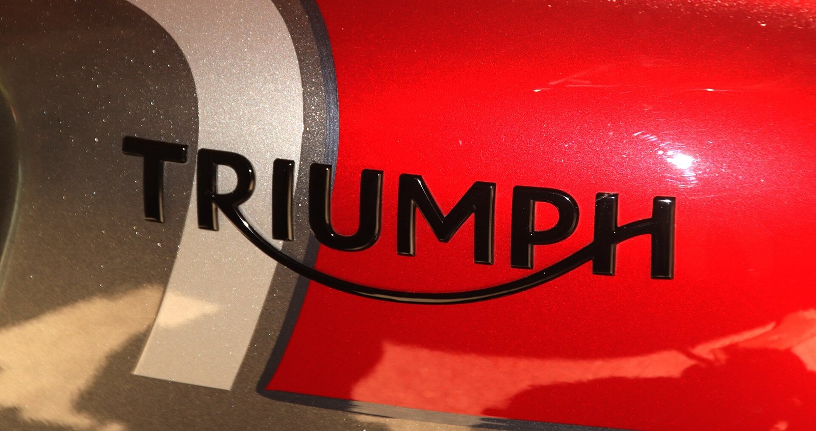 The current sans-serif TRIUMPH wordmark with its smile line, in black on a red and silver tank